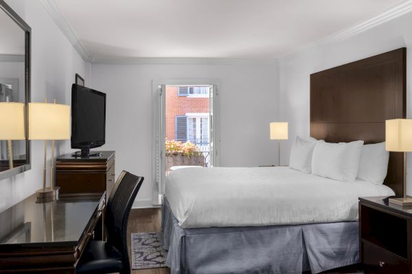 A neatly arranged hotel room features a bed, desk, chair, lamps, TV, and an open balcony door with a view of a brick building.