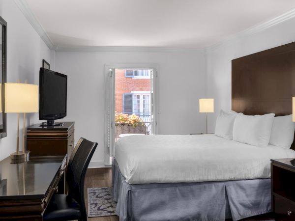 A neatly arranged hotel room features a bed, desk, chair, lamps, TV, and an open balcony door with a view of a brick building.