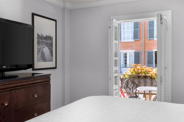 A room with a TV, framed photo, bed, and open doors leading to a small balcony with flowers and chairs, facing a building with shutters.