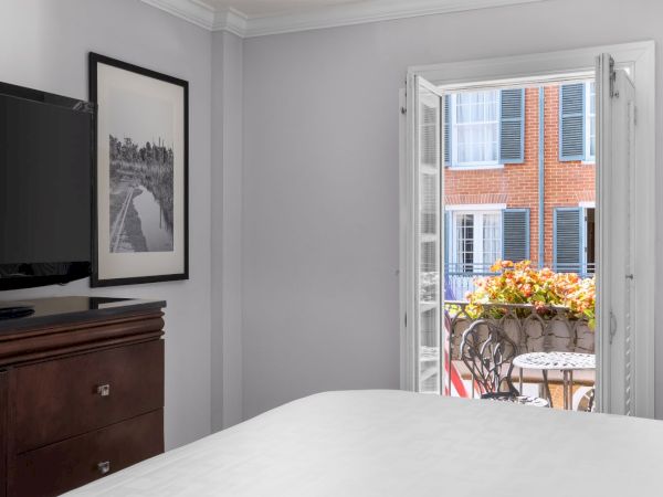 A room with a TV, framed photo, bed, and open doors leading to a small balcony with flowers and chairs, facing a building with shutters.
