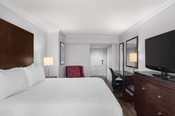 A hotel room with a bed, red chair, desk, lamp, and TV. The door is at the end of the room, and there's a large mirror on the wall.