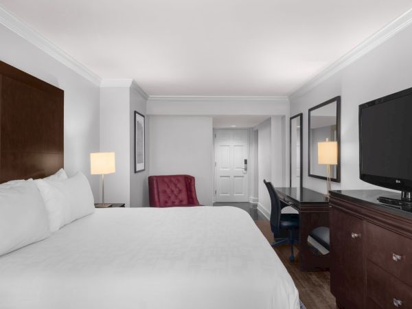 A hotel room with a bed, red chair, desk, lamp, and TV. The door is at the end of the room, and there's a large mirror on the wall.