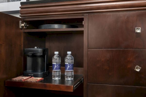 The image shows an open cabinet with a coffee maker, two bottled waters, and two coffee packets on a pull-out shelf.