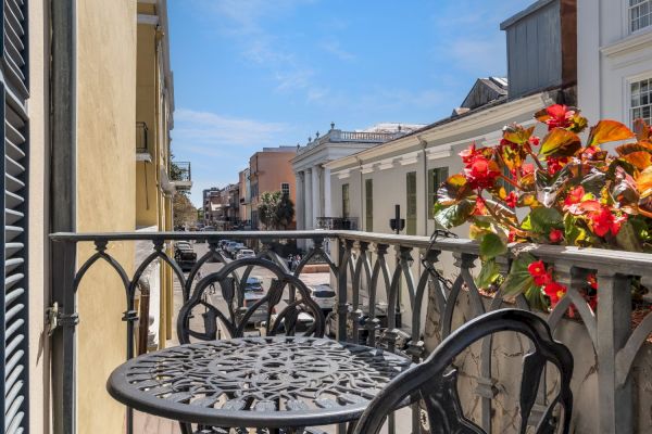 A quaint balcony with intricate metal chairs, a vibrant flower pot, overlooks a charming street with classic buildings on a sunny day.