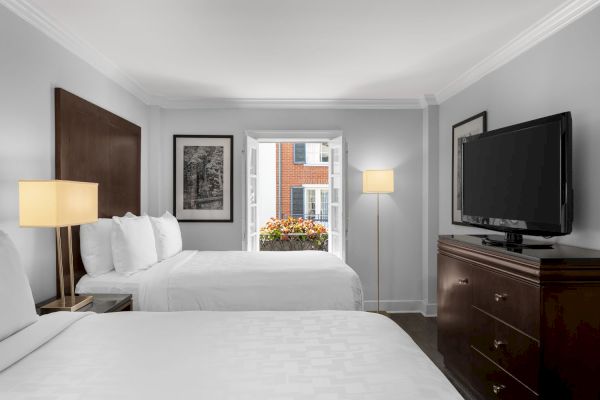 The image shows a hotel room with two beds, lamps, a dresser with a TV, and an open window with flowers and a view of a brick building.