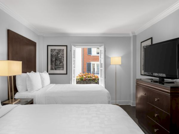 The image shows a hotel room with two beds, lamps, a dresser with a TV, and an open window with flowers and a view of a brick building.