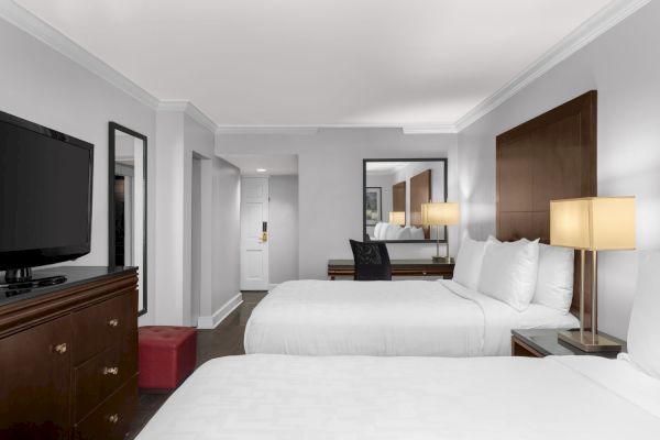 A hotel room with two beds, a dresser with a TV, a desk, lamps, a mirror, and a red ottoman near the room entrance.