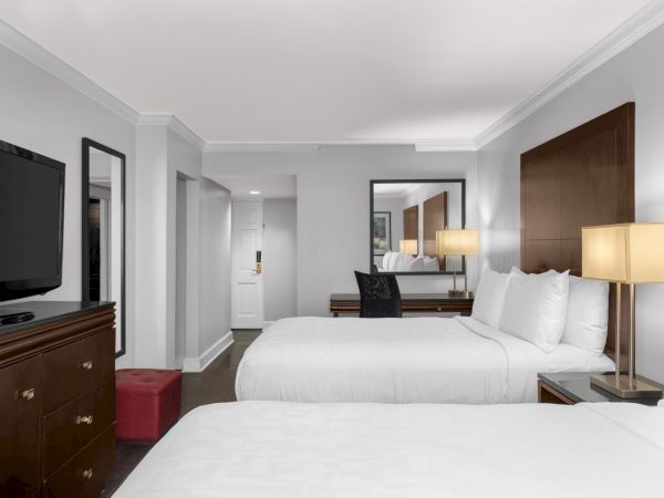 A hotel room with two beds, a dresser with a TV, a desk, lamps, a mirror, and a red ottoman near the room entrance.