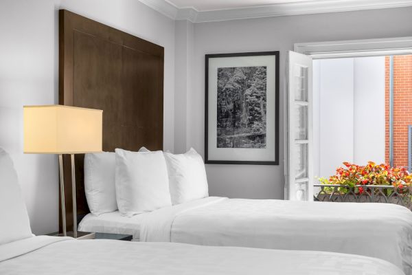 A hotel room with two white beds, a bedside lamp, framed nature art, and a window with blooming flowers outside.