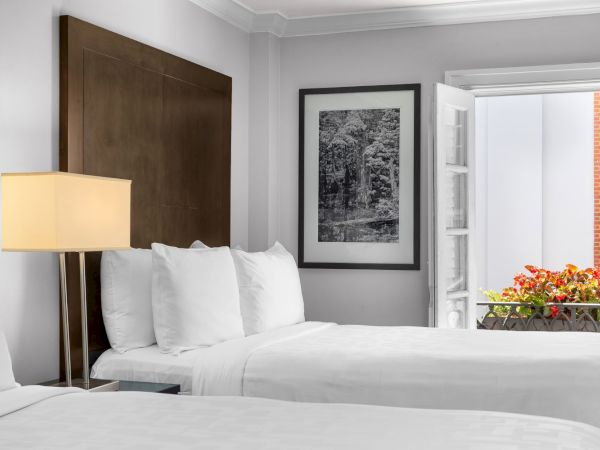 A hotel room with two white beds, a bedside lamp, framed nature art, and a window with blooming flowers outside.