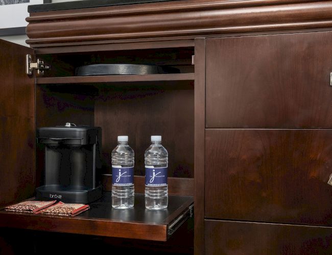 A dark wooden cabinet with an open drawershowing two bottled water, a coffee maker, a small plate, and a box inside the drawer.