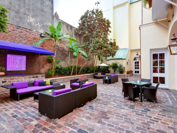 A stylish outdoor lounge with purple-cushioned seating, brick-paved courtyard, palm trees, and a small dining area in a charming courtyard.