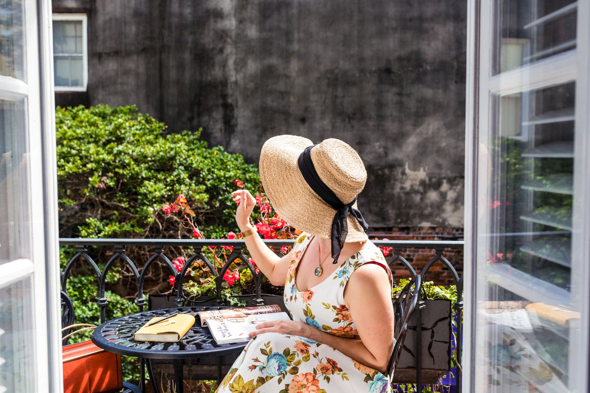 A woman in a floral dress and sunhat sits at an outdoor table with a book, enjoying a garden view through open doors.