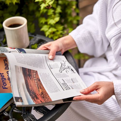 A person in a white robe is reading a magazine with a coffee cup nearby, on a table.