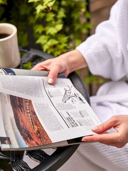 A person in a white robe is reading a magazine with a coffee cup nearby, on a table.
