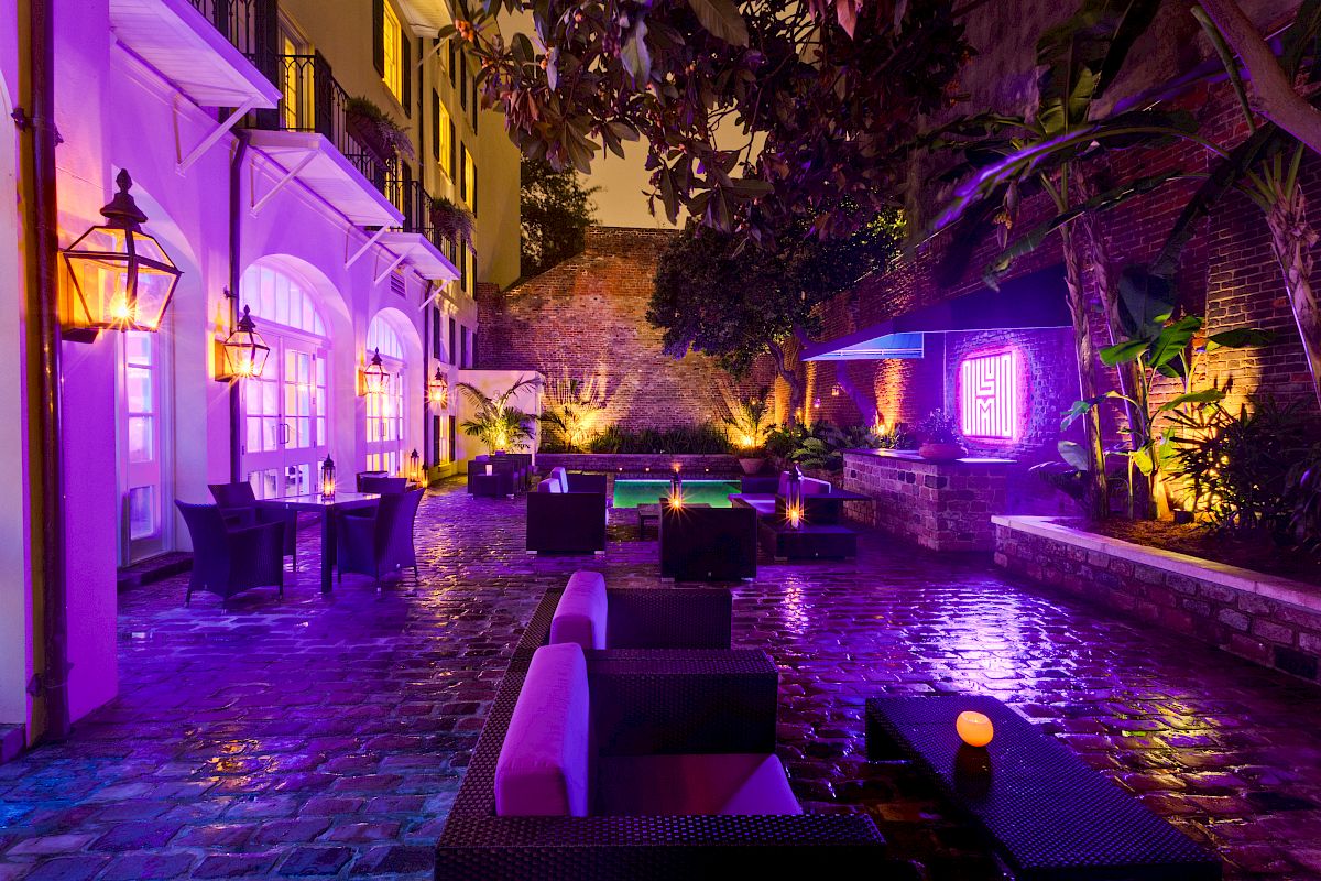 A dimly lit, vibrant outdoor courtyard with purple lighting, modern seating, and lush greenery.