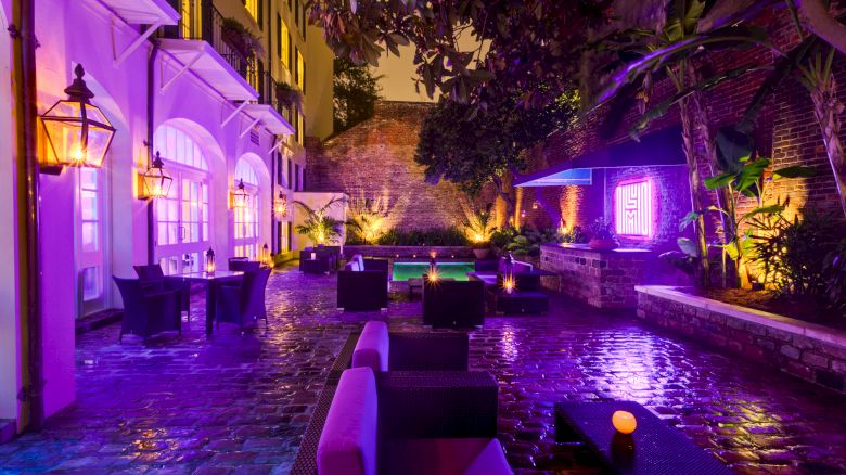 A dimly lit, vibrant outdoor courtyard with purple lighting, modern seating, and lush greenery.