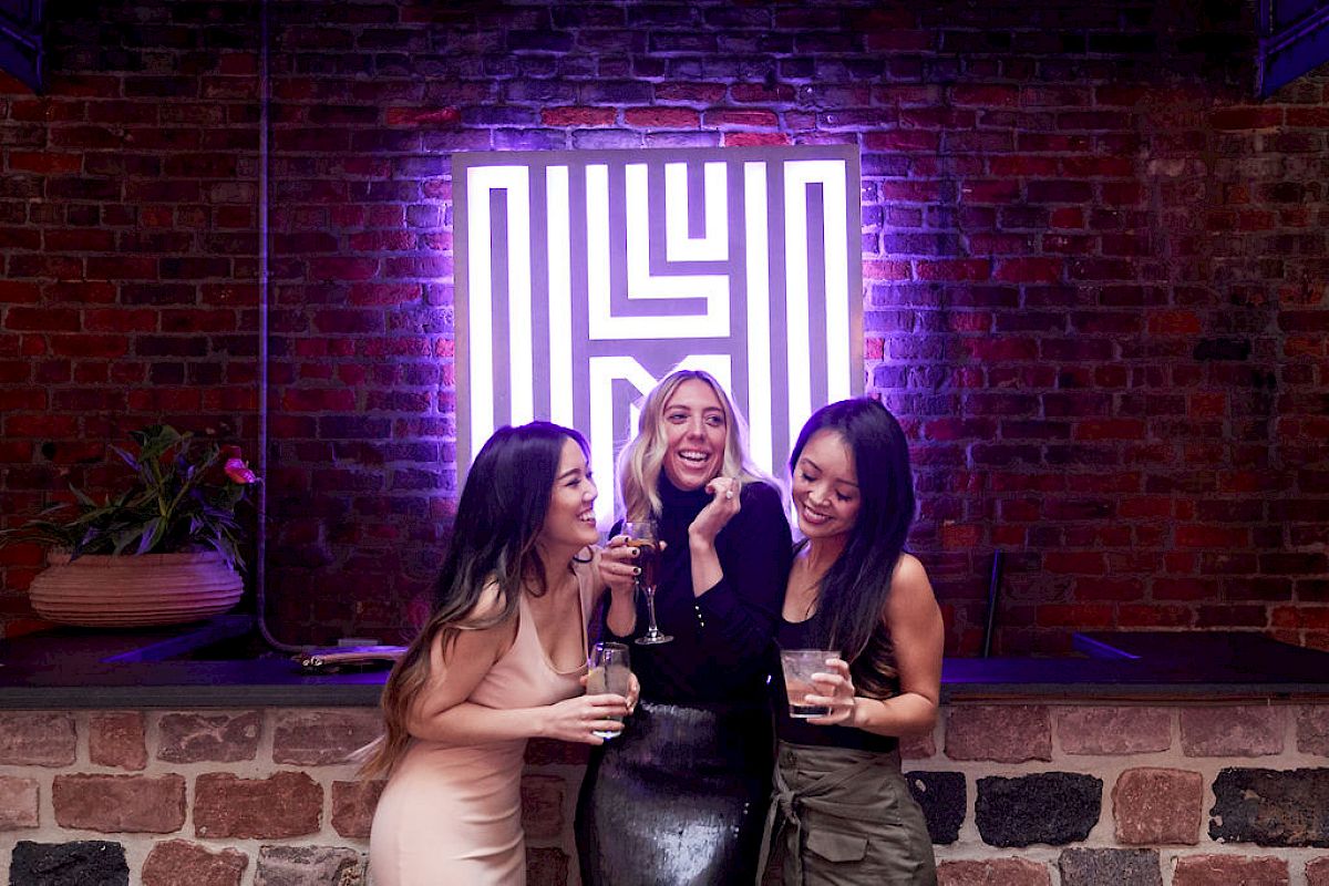 Three people are smiling and holding drinks in front of a neon sign on a brick wall.