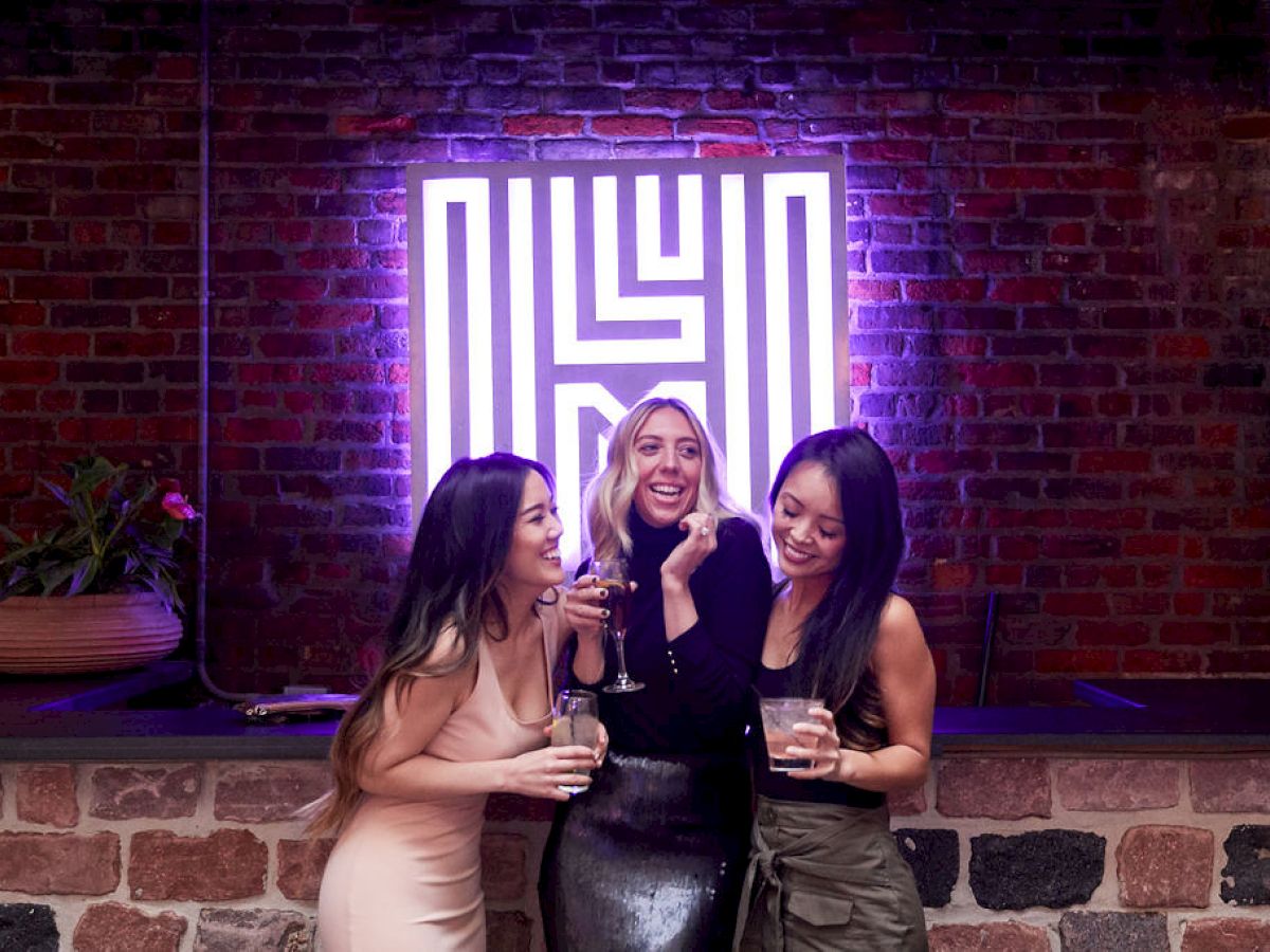 Three people are smiling and holding drinks in front of a neon sign on a brick wall.