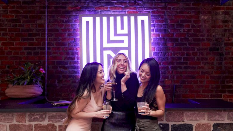 Three people are smiling and holding drinks in front of a neon sign on a brick wall.