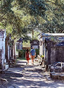 A couple walks through an old cemetery with above-ground tombs, surrounded by trees and dappled sunlight filling the path.