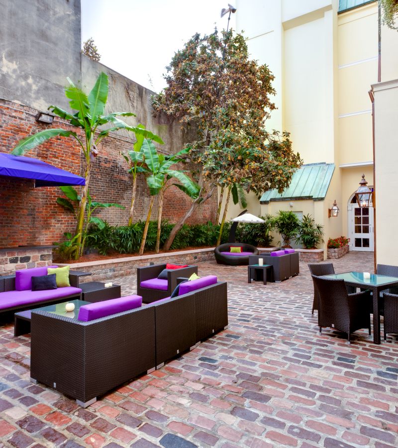 A cozy outdoor seating area with purple couches and chairs, surrounded by plants and a brick wall.