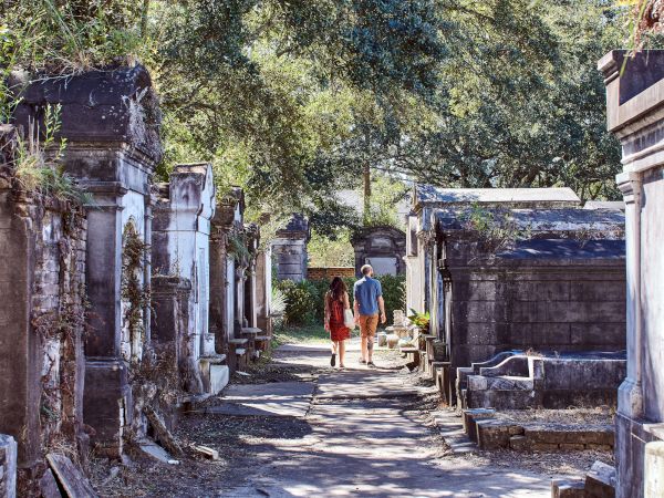 A couple walks through a historic cemetery, surrounded by large mausoleums and greenery, under the shade of trees lining the path.