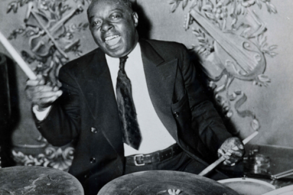 A man in a suit enthusiastically plays a drum set in front of an ornate backdrop with decorative musical instruments.