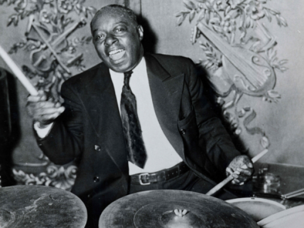A man in a suit enthusiastically plays a drum set in front of an ornate backdrop with decorative musical instruments.