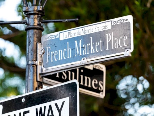 Street signs display "French Market Place" and "Ursulines" with a "One Way" sign below, amidst a background of greenery.