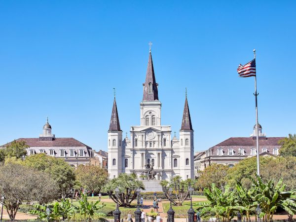 The image shows a historic cathedral with three spires surrounded by trees and buildings, and a U.S. flag waving in the foreground.