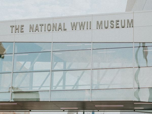 The image shows a walkway labeled "THE NATIONAL WWII MUSEUM" with a few people and a sculpture in the foreground.