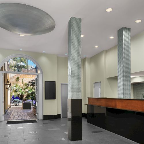 A modern lobby area with a high ceiling, sleek columns, a reception desk, and an open entrance leading to a lush outdoor patio area.