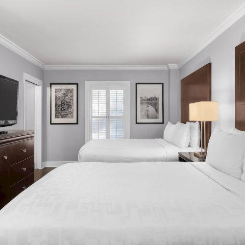 The image shows a hotel room with two beds, a TV on a wooden dresser, and framed pictures on a gray wall.