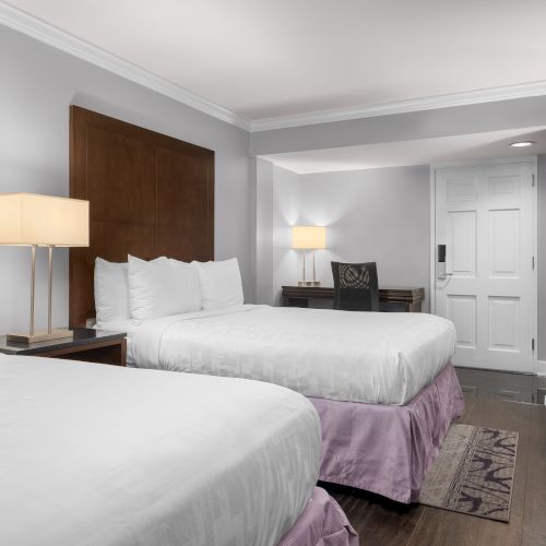The image shows a hotel room with two beds, a TV, lamps, a desk, and a chair. The room has a modern and clean design.