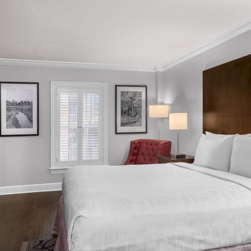 A tidy hotel room with a bed, red chair, TV, framed photos, lamps, and window shutters.