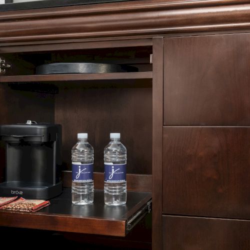 A wooden cabinet with a coffee maker, two water bottles, and coffee packets inside the open door.