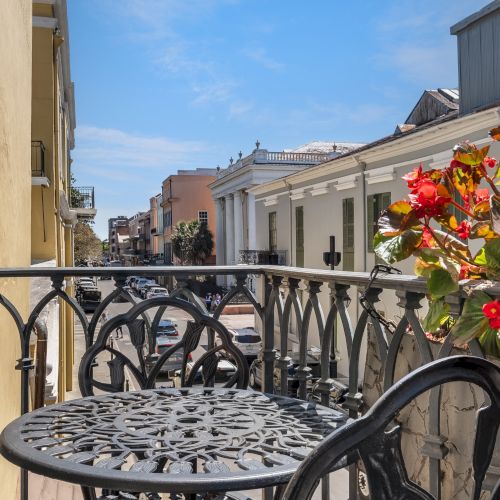 A balcony with an ornate metal table and chair overlooks a street lined with colorful buildings and flowers, under a clear blue sky.