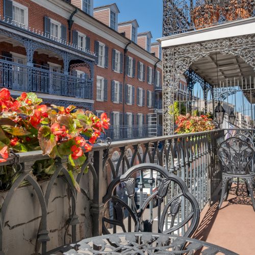 A balcony with ornate ironwork, vibrant red flowers, and outdoor seating overlooks a street with classic architecture.