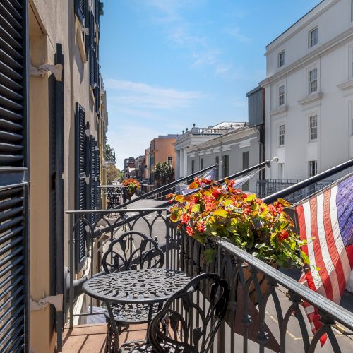 A balcony view overlooking a street with U.S. flags, wrought iron furniture, and colorful flowers, amidst historic buildings under a blue sky.