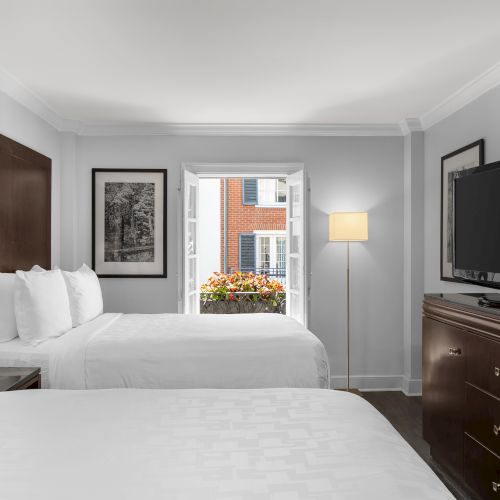 A hotel room with two beds, a TV, and a view of a building through open doors with flowers on the ledge.