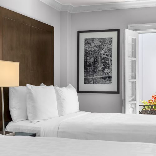A hotel room features two white beds, a bedside lamp, and a framed black and white photo, with flowers seen through an open window.
