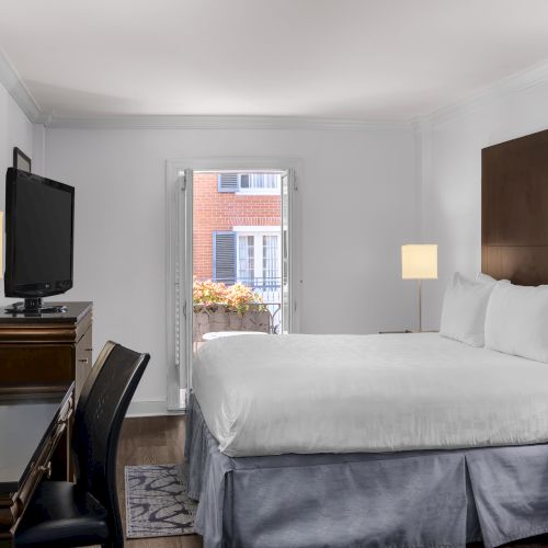 A cozy hotel room features a bed, desk, chair, lamps, TV, and an open window with flowers outside the view.