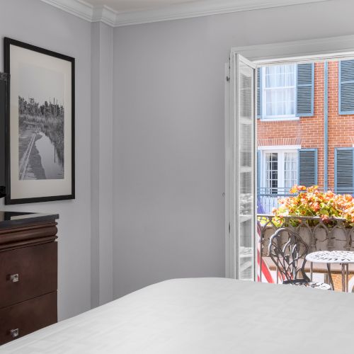 A cozy room with a TV, framed artwork, bed, and an open door to a balcony with a small table and flowers.