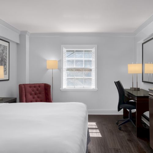 A modern hotel room with a bed, red chair, desk, lamp, TV, and window that lets in natural light, creating a cozy space.