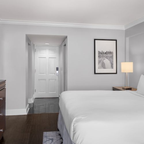 A hotel room with a bed, TV, lamp, and artwork on the wall, with a door leading to an entrance.