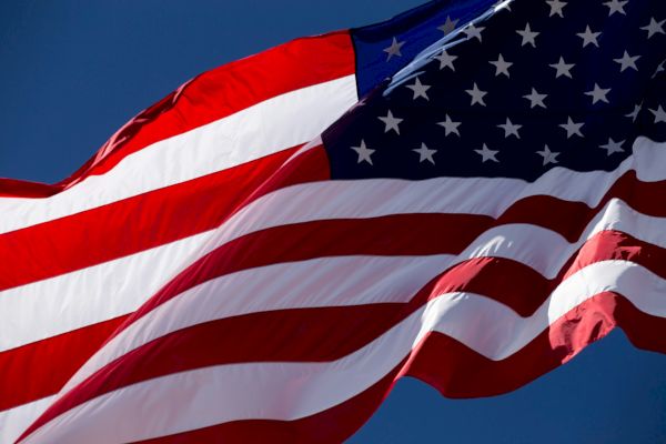 The image shows the United States flag waving against a clear blue sky, displaying its red, white, and blue colors with stars and stripes.