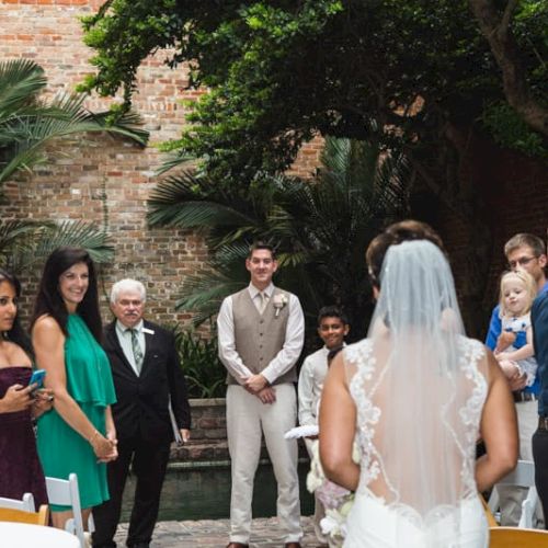 A bride faces a group of people dressed formally, possibly at an outdoor wedding or event, with lush greenery and brick walls.
