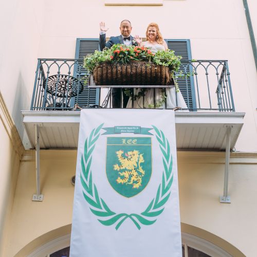 A couple stands on a balcony with a crest banner hanging below. The banner features a lion emblem surrounded by a wreath design.