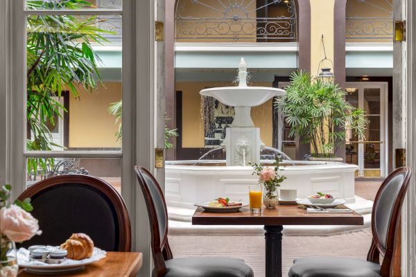 A charming courtyard with a central fountain, viewed from a dining area with tables set for breakfast, including juice and pastries.